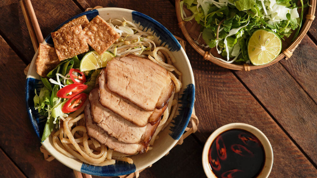 A bowl of Cao Lau noodles with slices of pork, greens, crunchy croutons and thick white noodles in a light broth